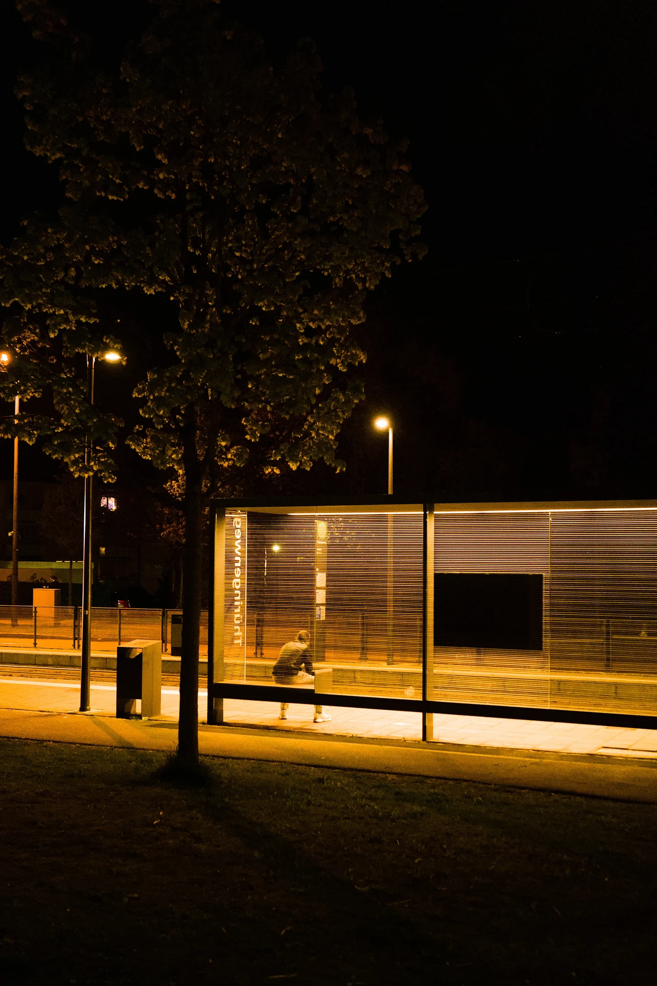 A person sitting lonely at a tram stop at night. The scene stop is well-lit, but the light quickly fades into darkness.