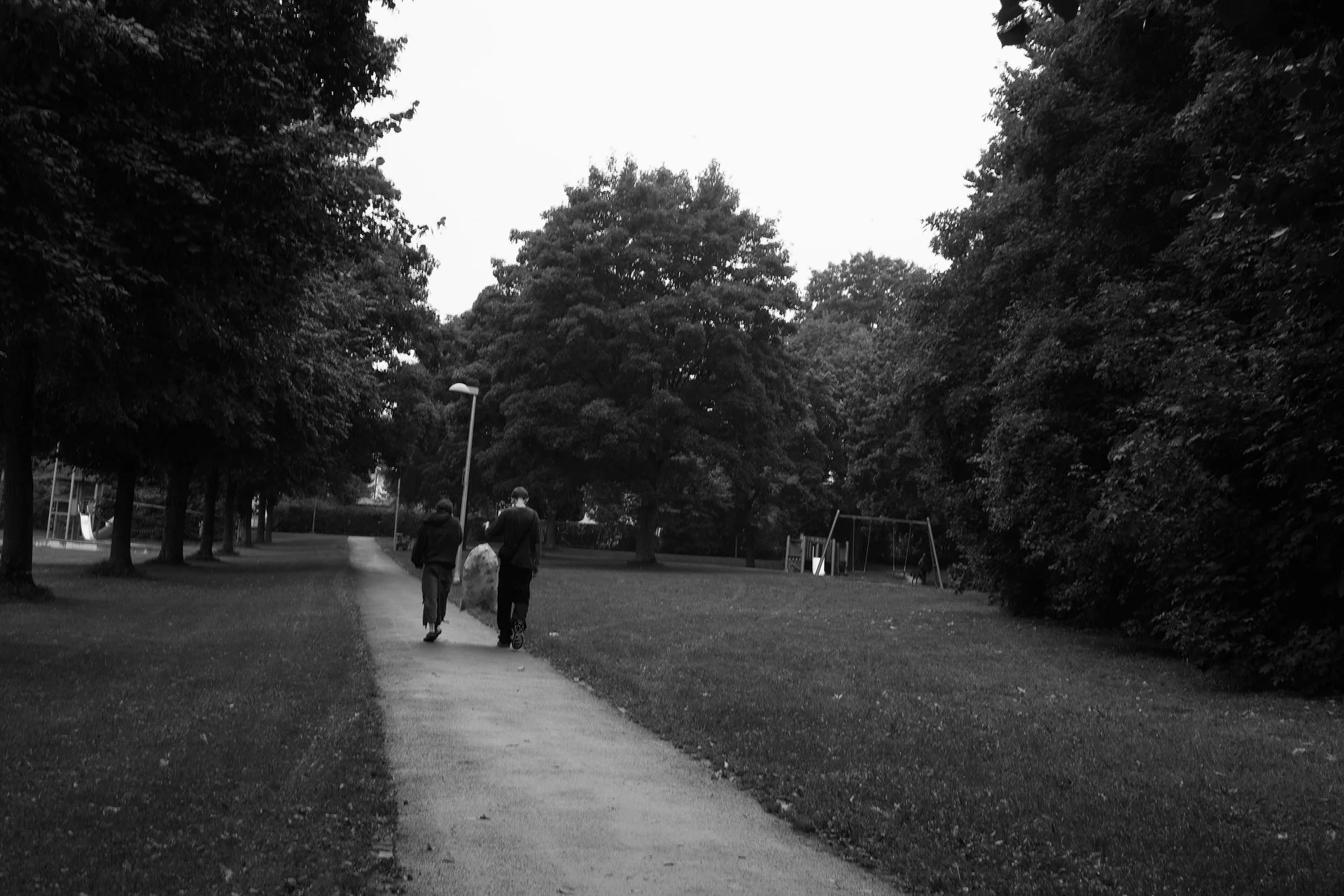 Two person walking away on a pedestrian way in a park. Together the carry a large bin bag.
