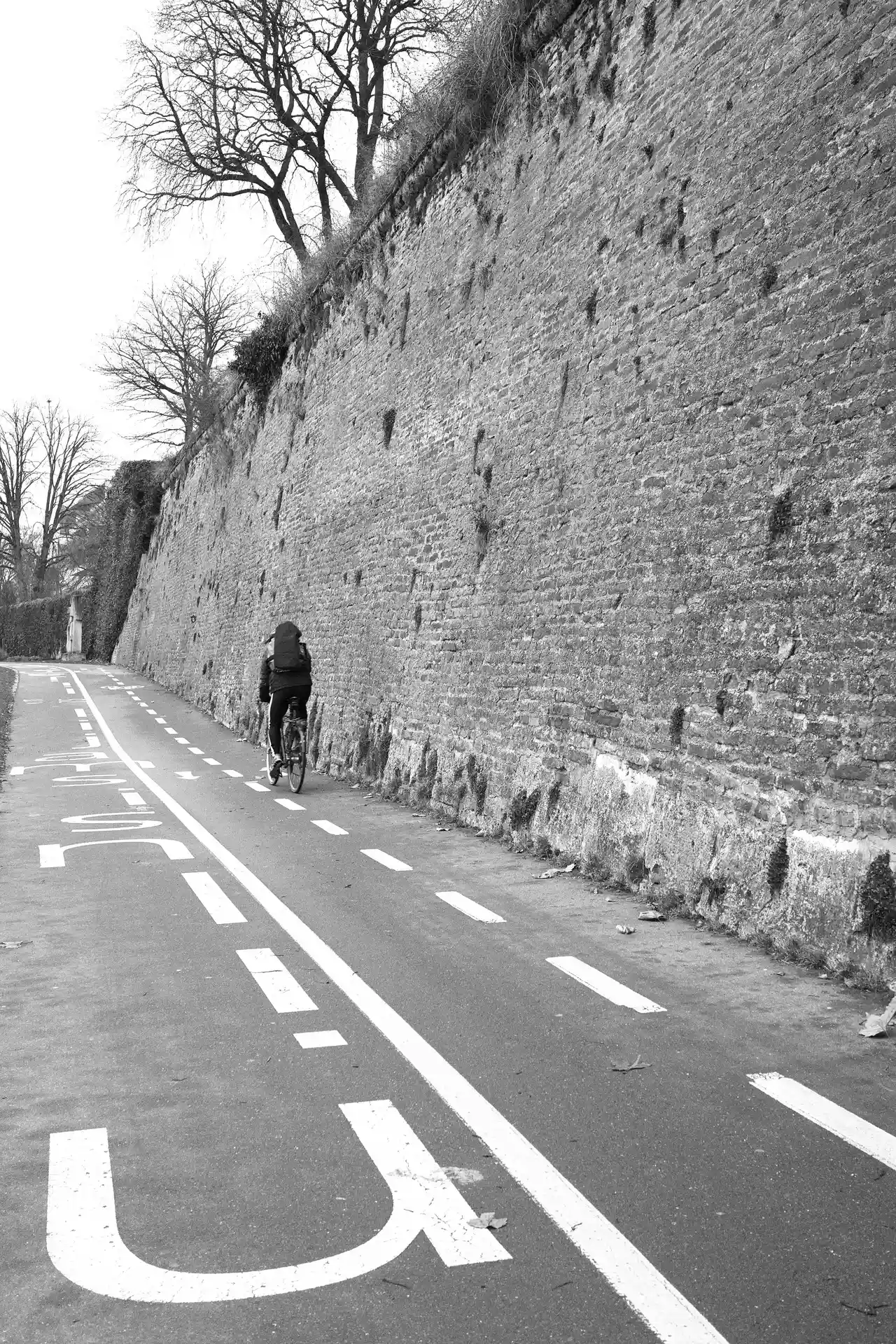 A black and white photo. On the right side is the city wall of Ulm, in the center and on the left is a pedestrian and cycle way. A person is currently cycling away from the photographer.