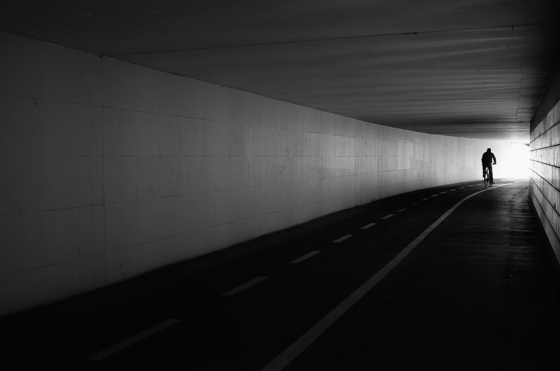 A subway for cycilst and pedestrians. Only a single cyclist is in the frame at the end of the subway, contrasted against the outside light.