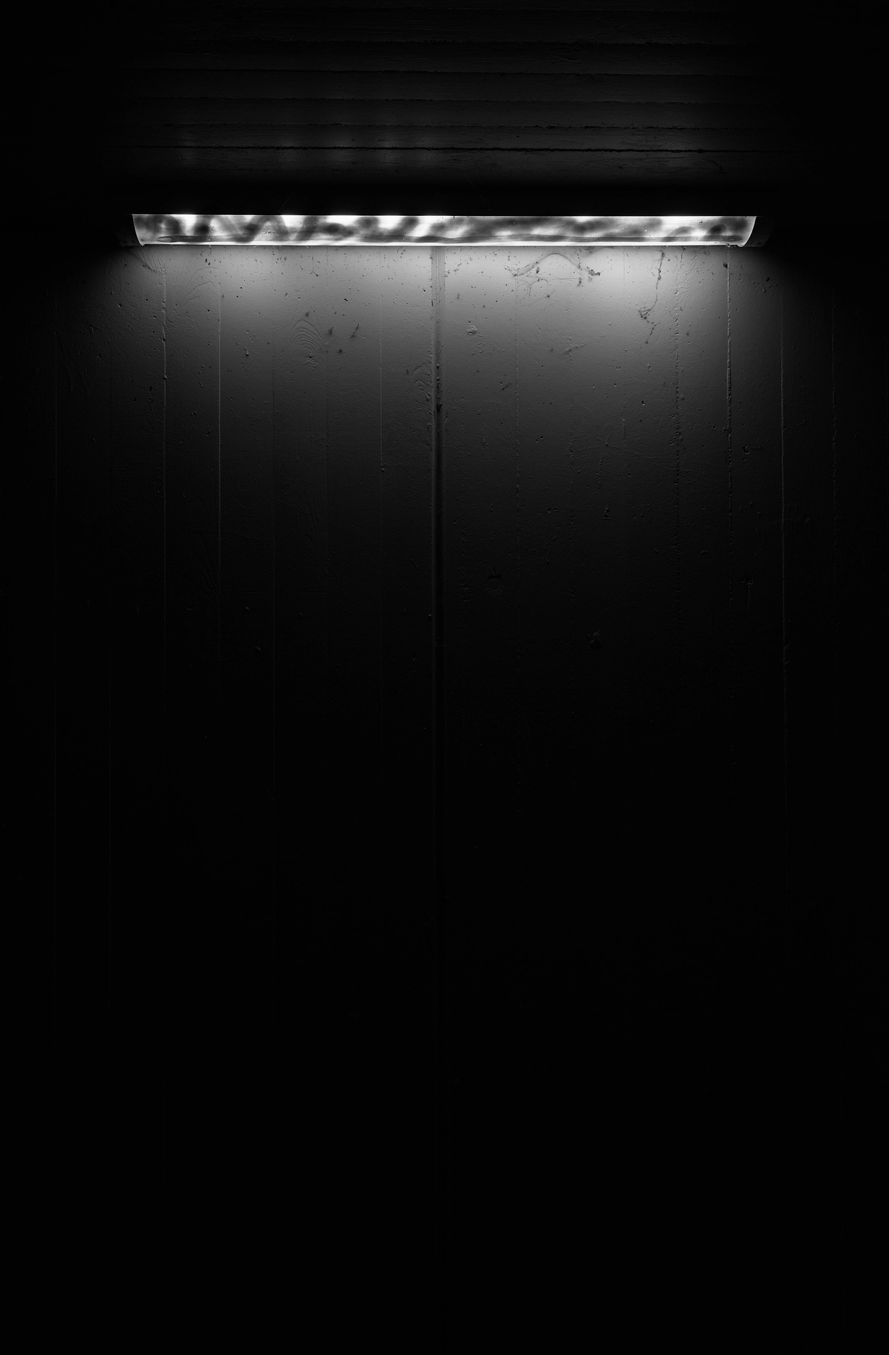 A ceiling light in front of a concrete wall. The light is quite dimm and everything quickly fades into black.