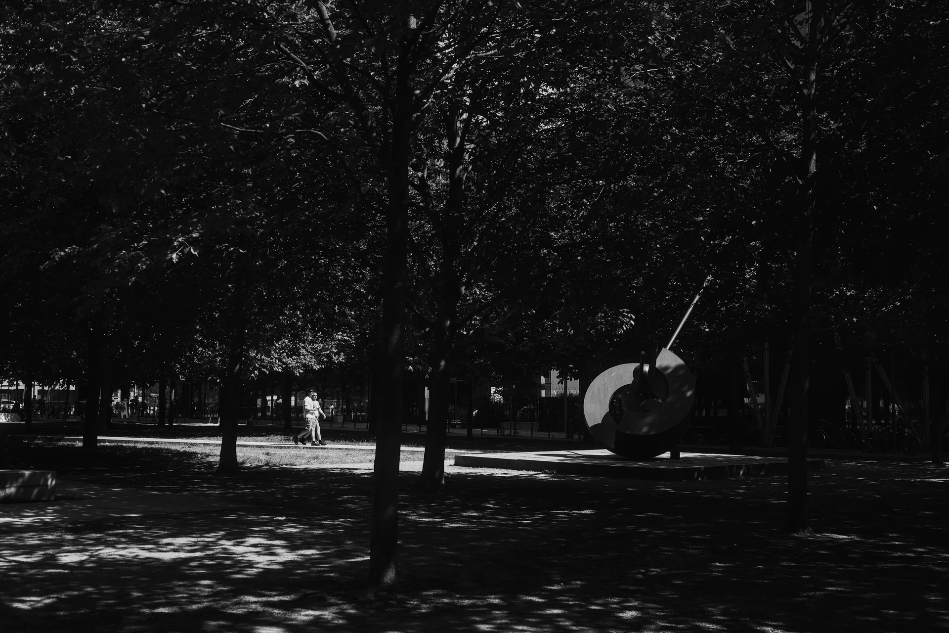 Black and white photo of a sculpture in a park. Framed by some tree trunks and highlighted by sunlight are two people walking towards it.