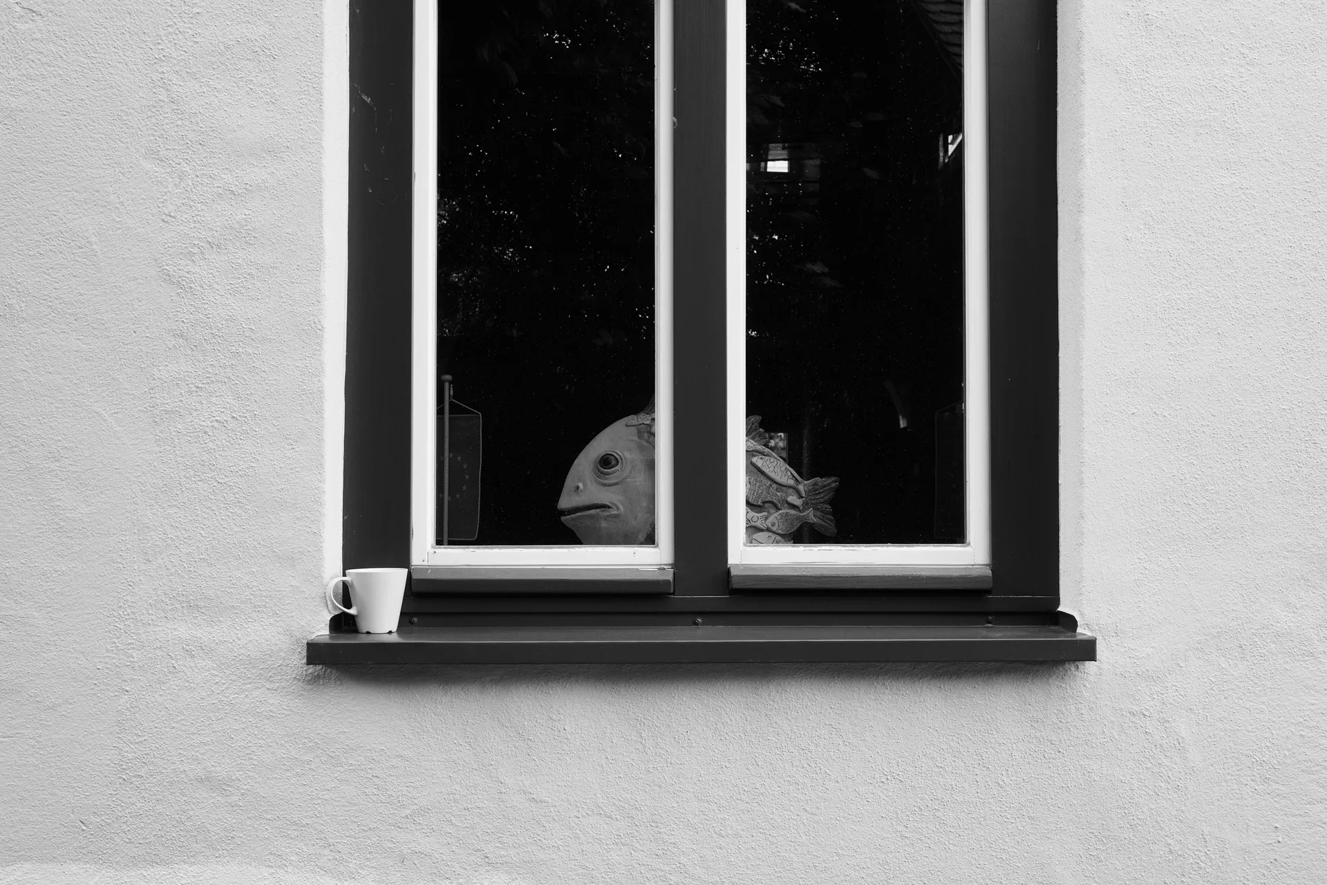 Black and white photo of a window frame with a cup on the windowsill and a sculpture of a fish on the inside.