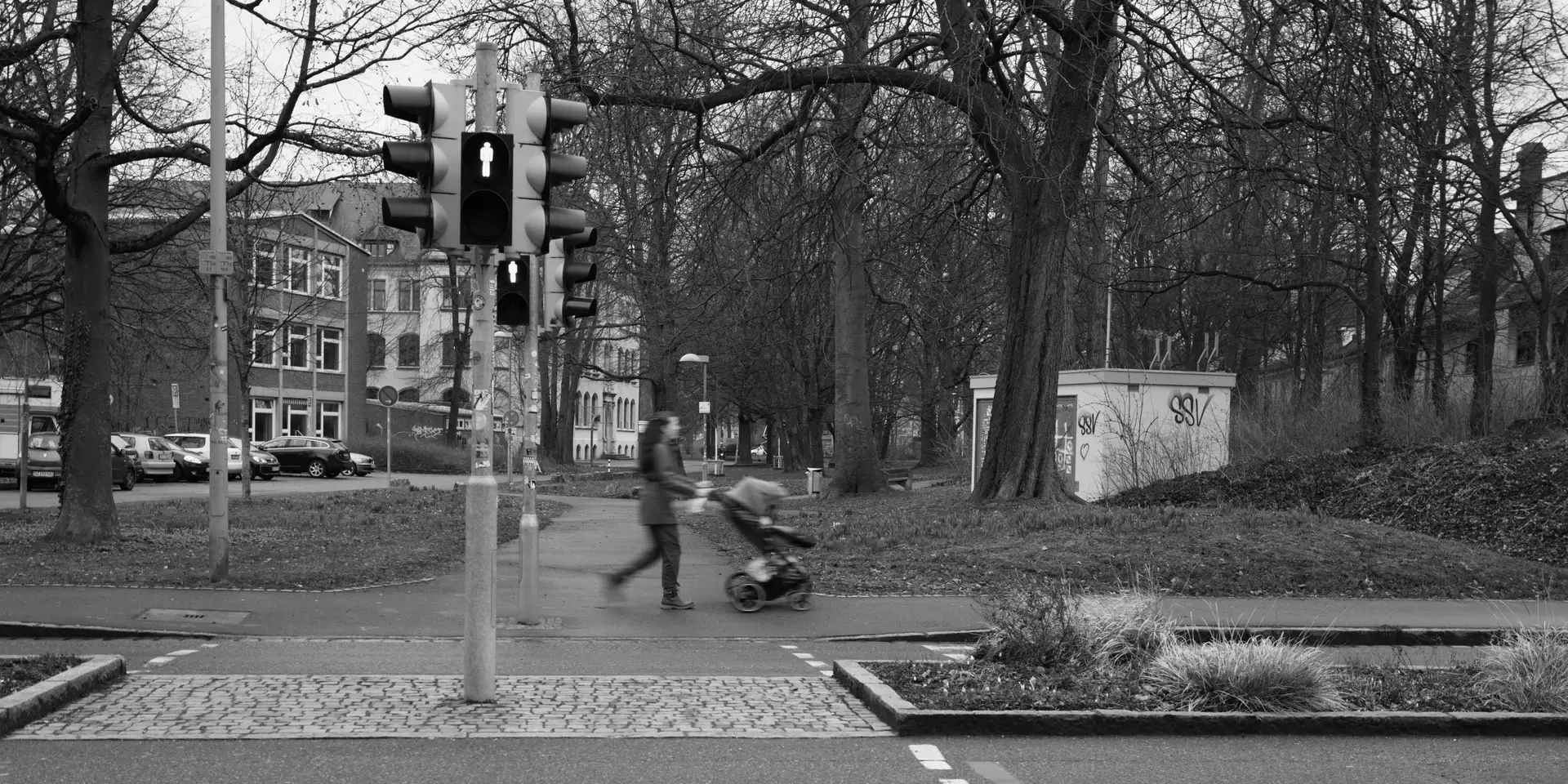 Picture of a pedestrian crossing with red light. On the opposite side a person walks by with a pram, blurred slightly due to the slow shutter speed.