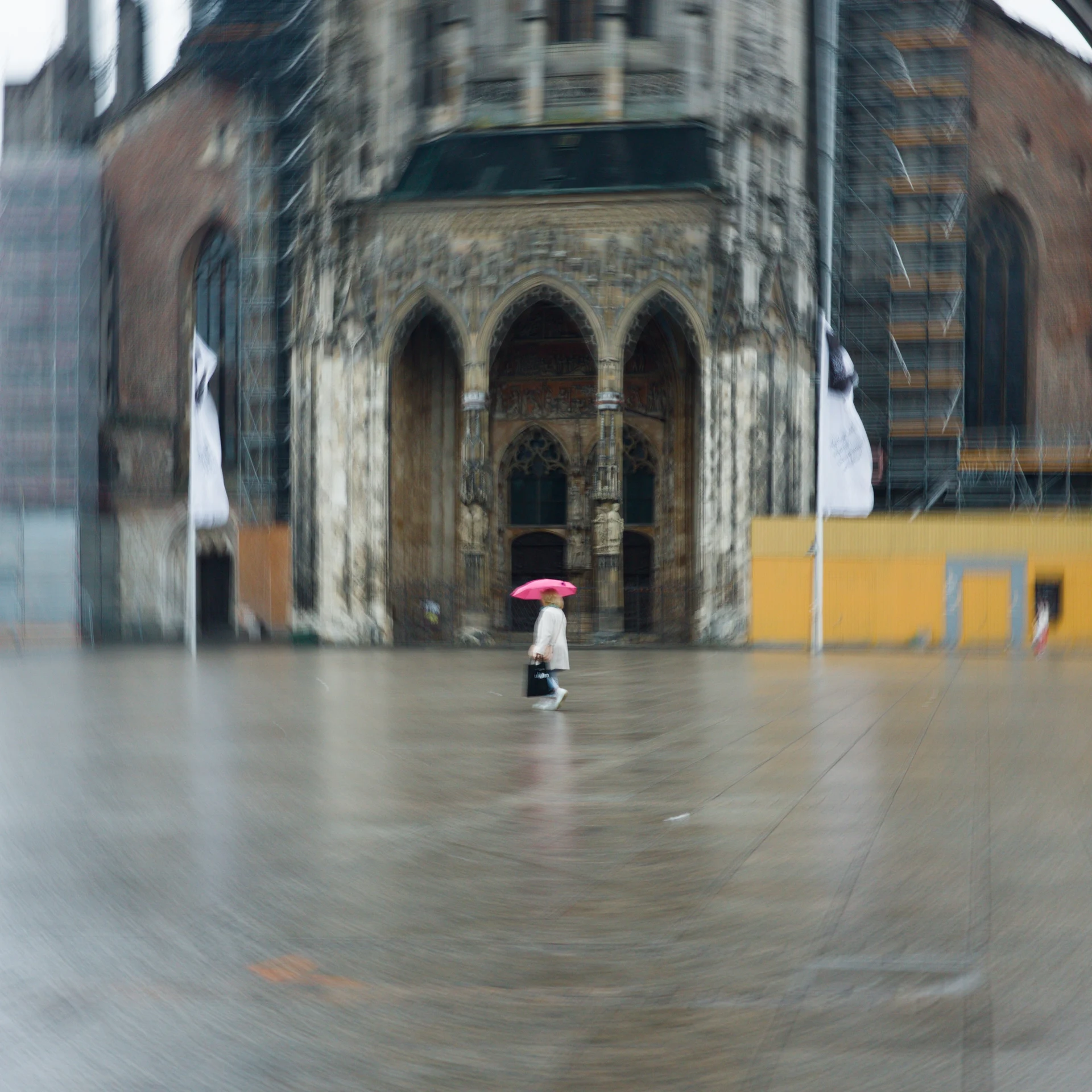 Picture of a person with a white jacket and a pink umbrella on the place in front of the Ulmer Münster during rain.  The camera was rotated during exposure resulting in a circular effect.